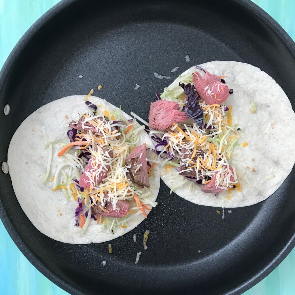 Tortillas open on a skillet with cheese, steak and veggies inside.