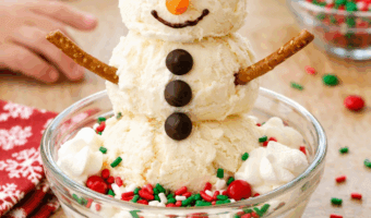 Homemade snowman ice cream sundae made with vanilla ice cream scoops, chocolate chip eyes, candy nose, and pretzel arms in a festive winter kitchen setting.