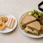Two white plates on a light counter: one with a turkey sandwich and a side salad, the other with hummus, crackers, and rolled deli meat, plus a blue water bottle nearby.