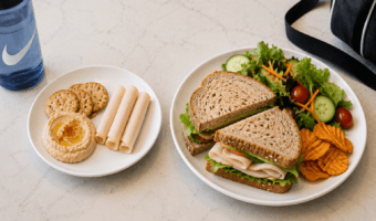 Two white plates on a light counter: one with a turkey sandwich and a side salad, the other with hummus, crackers, and rolled deli meat, plus a blue water bottle nearby.